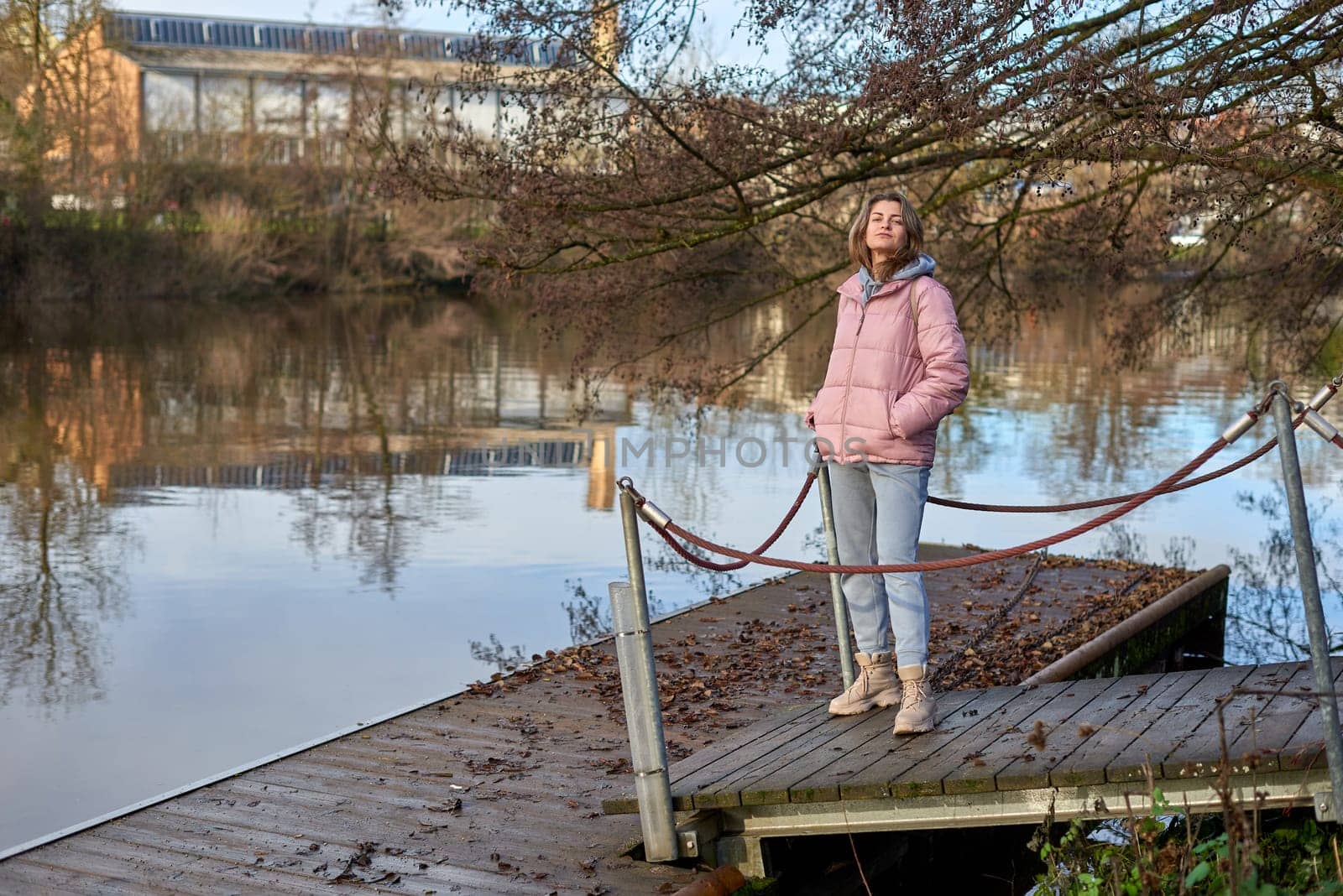 A young woman standing at the shore looking at the river in autumn sunny day. Street view, copy space for text, travel photo. Happy tourist woman on the bank of the river in autumn in warm clothes. Tourists enjoy their vacation, winter season. Romantic look and travel concept. A joyful mood in a caucasian girl. Winter Wonderland: Enchanting Girl by the Riverside in Autumn by Andrii_Ko