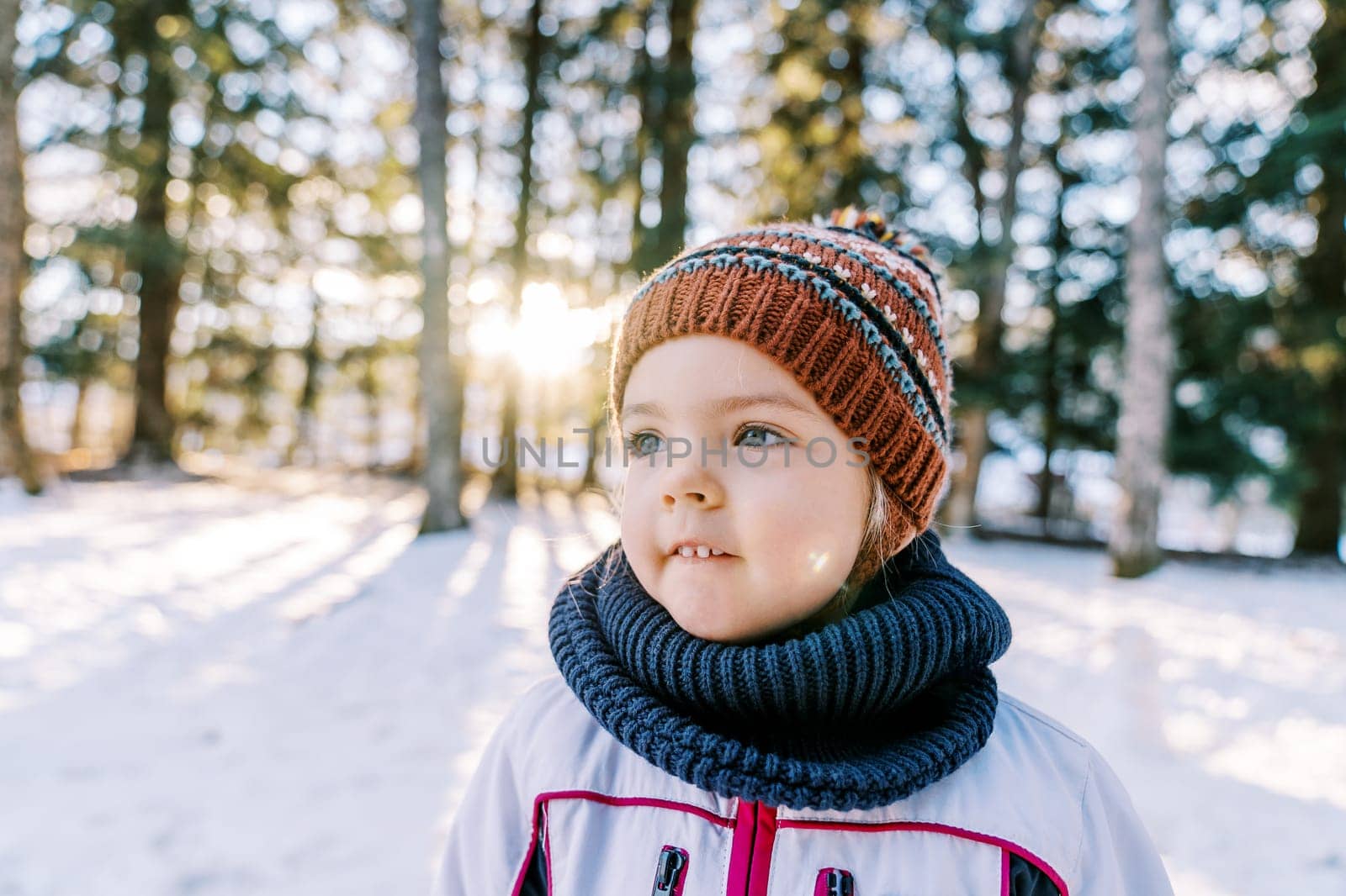 Little smiling girl in a hat and scarf stands in a snowy forest and looks away. High quality photo