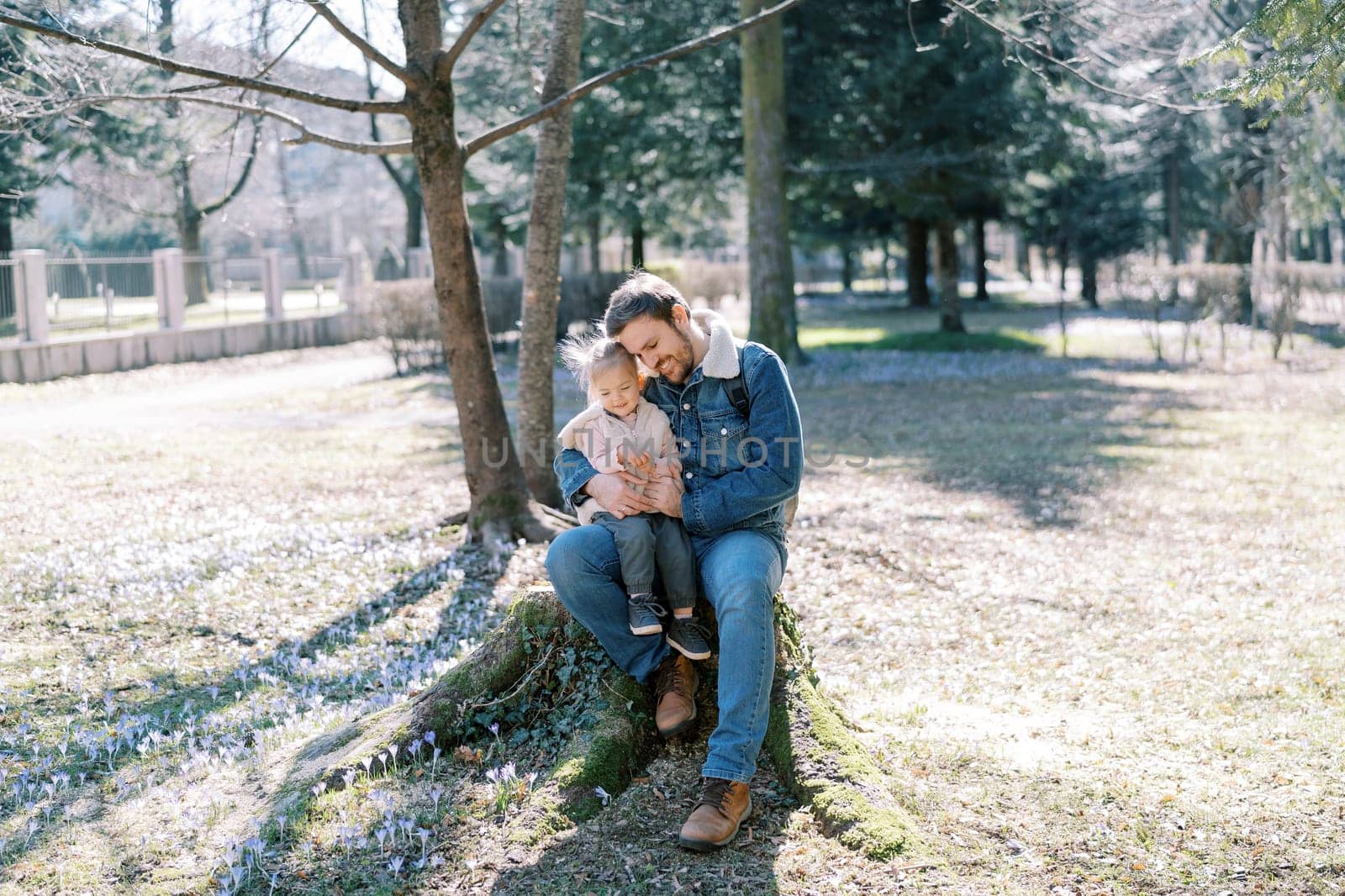 Smiling dad touches with his head the head of a little girl sitting on his knees on a stump in the park. High quality photo