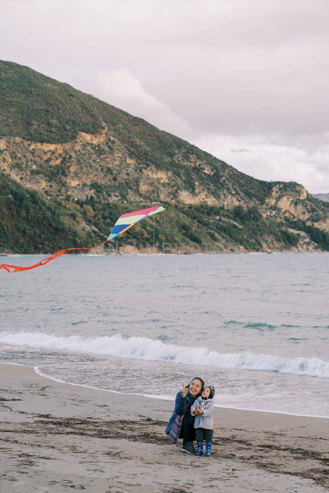 Mom sat down near her little daughter, showing her a kite hovering above. High quality photo