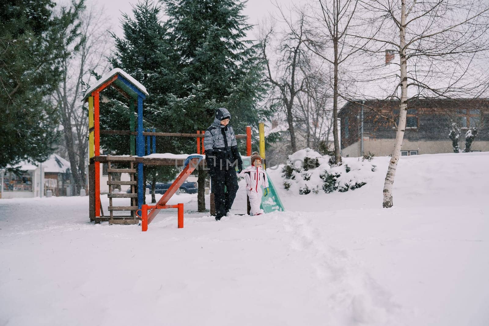 Little girl and her mother walk holding hands from a colorful snow-covered slide through the park under snowfall by Nadtochiy