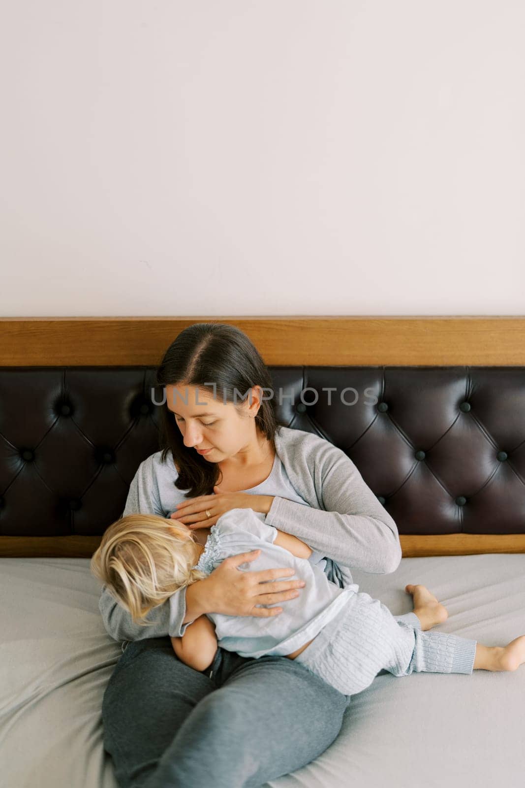 Mom breastfeeds a little girl hugging her in her arms while sitting on the bed by Nadtochiy