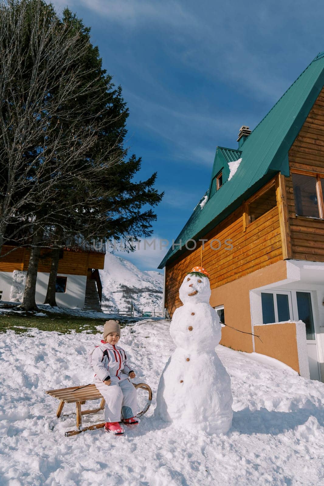 Little girl sits on a sleigh near a snowman in the courtyard of a wooden chalet by Nadtochiy