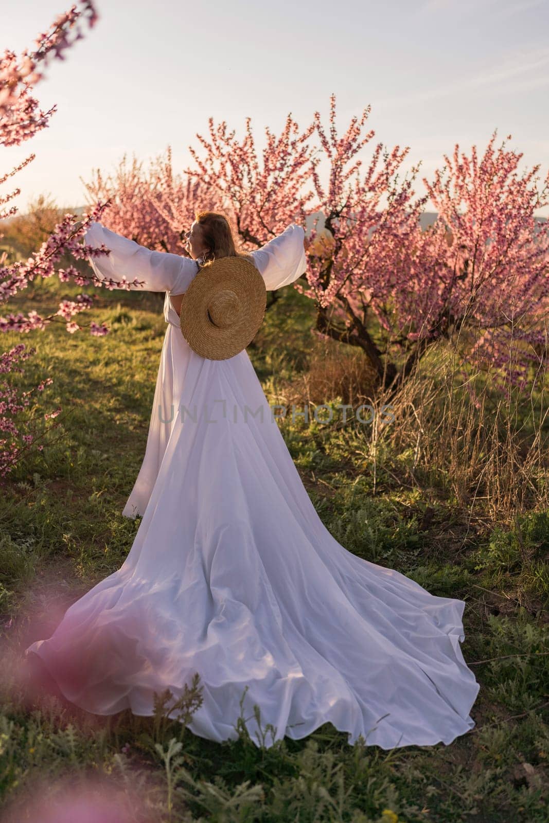 Woman blooming peach orchard. Against the backdrop of a picturesque peach orchard, a woman in a long white dress and hat enjoys a peaceful walk in the park, surrounded by the beauty of nature