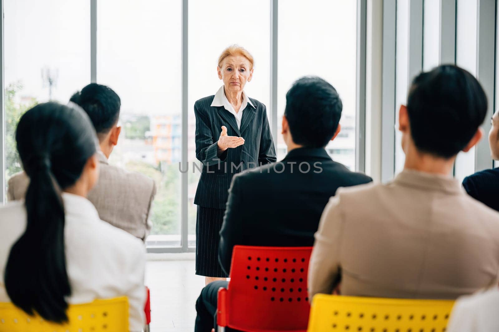 A confident senior corporate coach leads a seminar for a business team. The female director and CEO engage with employees in an office meeting, discussing projects and providing instructions.