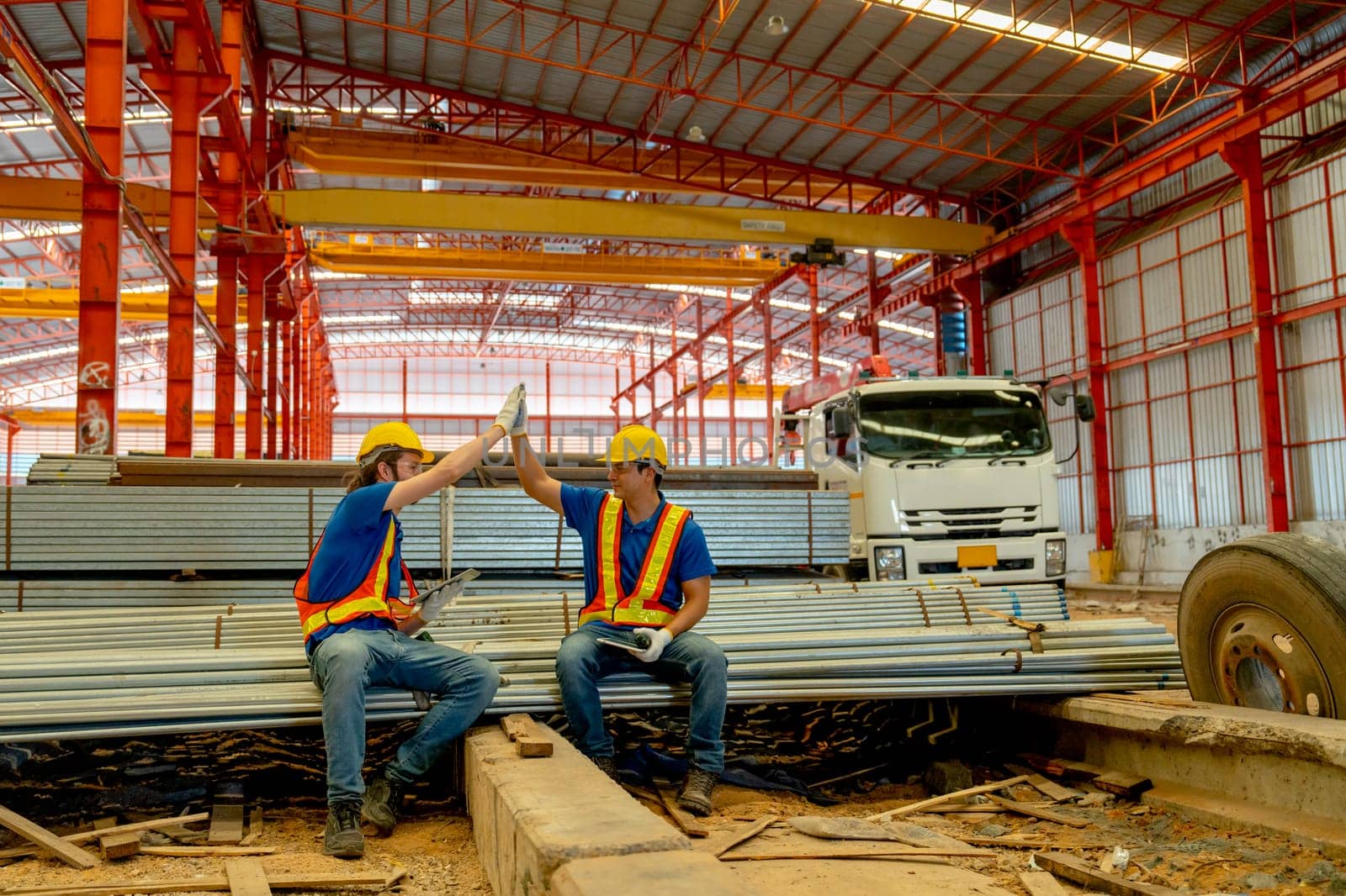Caucasian technician workers sit down on stack of metal pipe and do touch hands and look happy in factory workplace area.