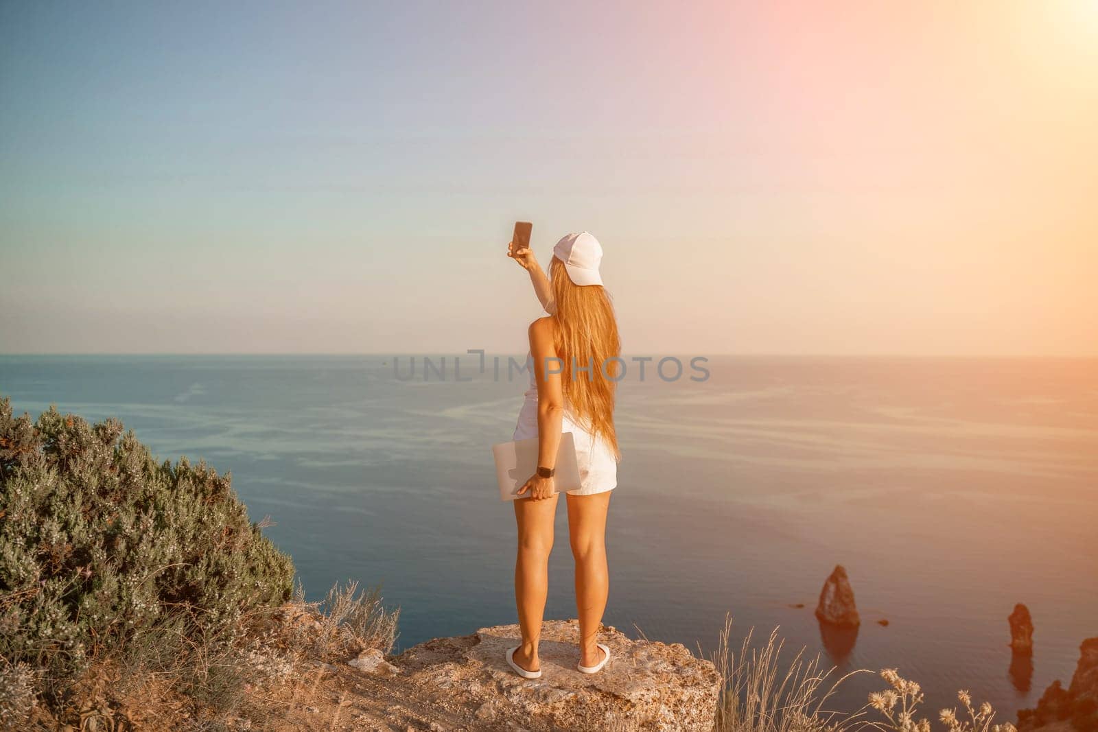 Selfie woman sea. The picture depicts a woman in a cap and tank top, taking a selfie shot with her mobile phone, showcasing her happy and carefree vacation mood against the beautiful sea background by Matiunina