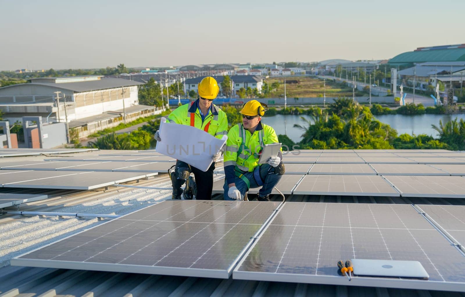 Front view of two Caucasian technician workers use construction plan and tablet to check and maintenance solar cell panels on rooftop of factory or the building.