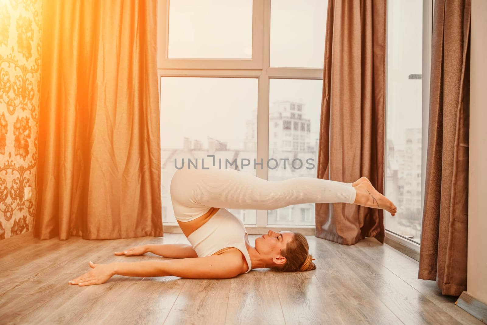 Young athletic attractive woman practicing yoga. Works out at home or in a yoga studio, sportswear, white pants and a full-length top indoors. Healthy lifestyle concept by Matiunina