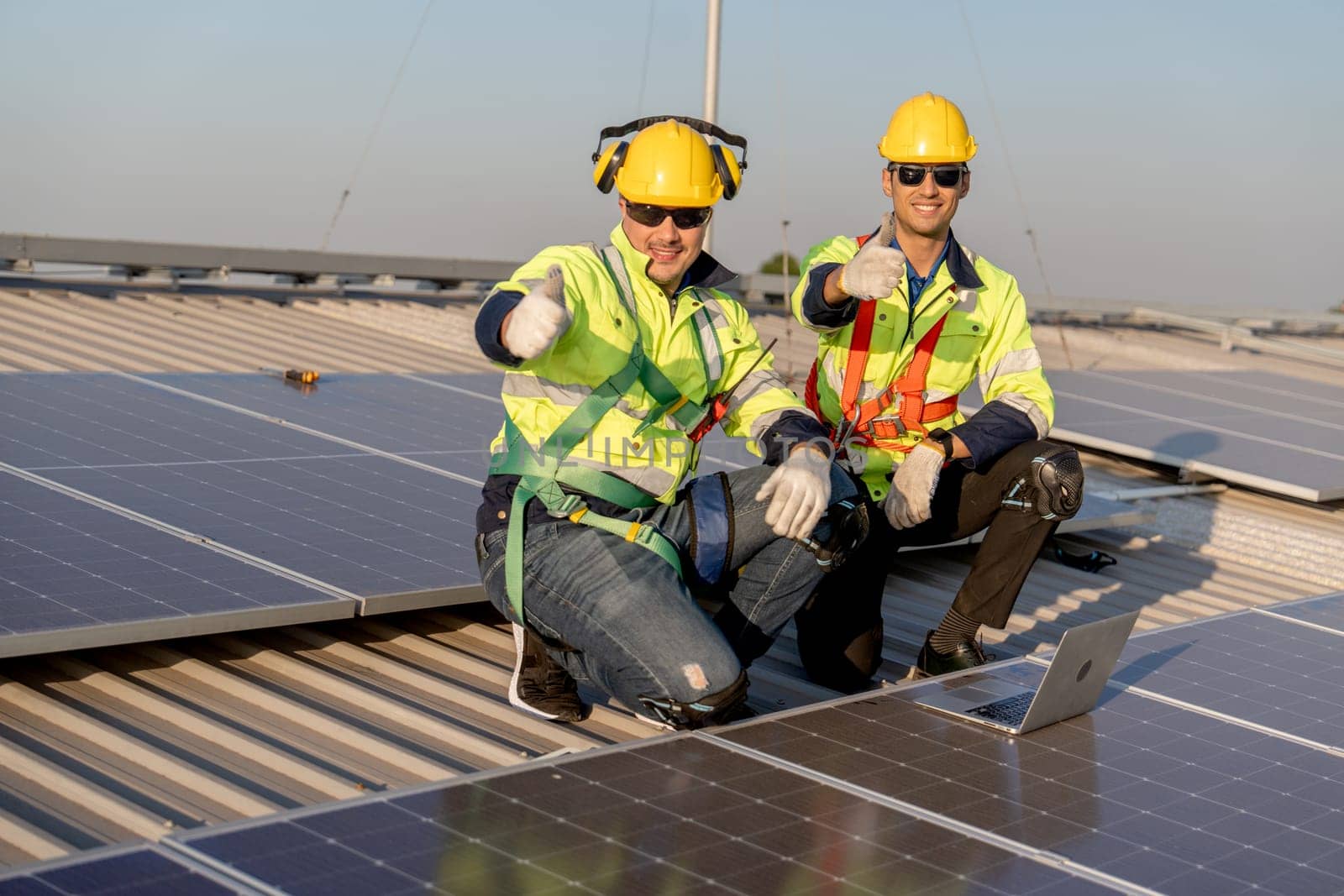 Two industrial technician worker sit near solar cell panels on rooftop of factory building and show thumbs up to camera with smiling. Green and sustainable energy concept.