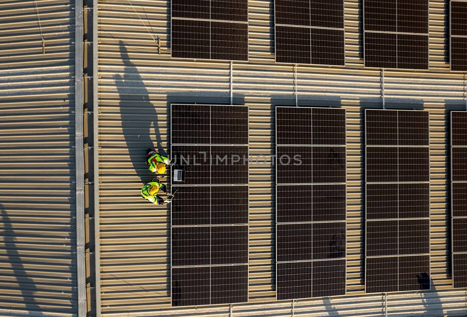 Vertical of drone view of two technician workers use laptop and tablet to check and maintenance solar cell panels on rooftop of factory building.