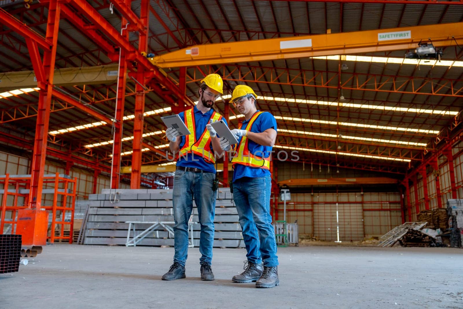 Wide shot of two Caucasian technician workers hold tablet and stand together to discuss about work in factory workplace area.