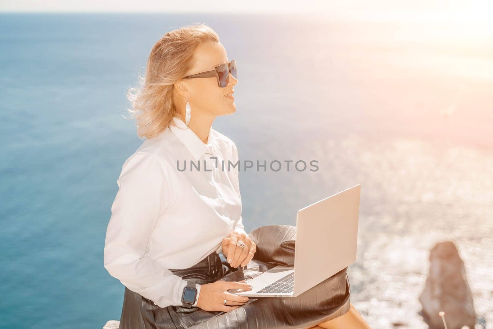 Business woman on nature in white shirt and black skirt. She works with an iPad in the open air with a beautiful view of the sea. The concept of remote work. by Matiunina