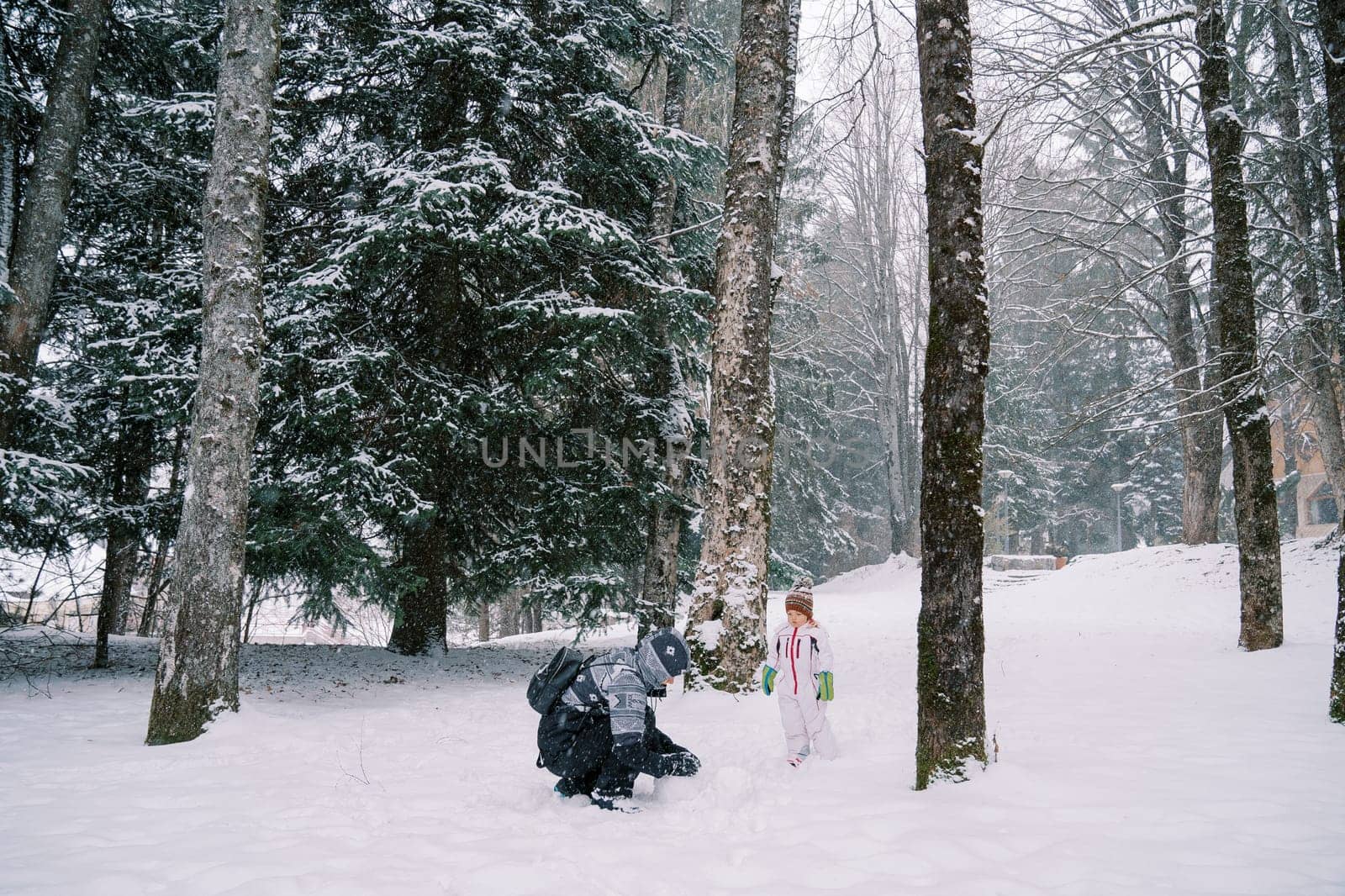 Little girl stands in a snowy forest and looks at her mom making a snowman by Nadtochiy