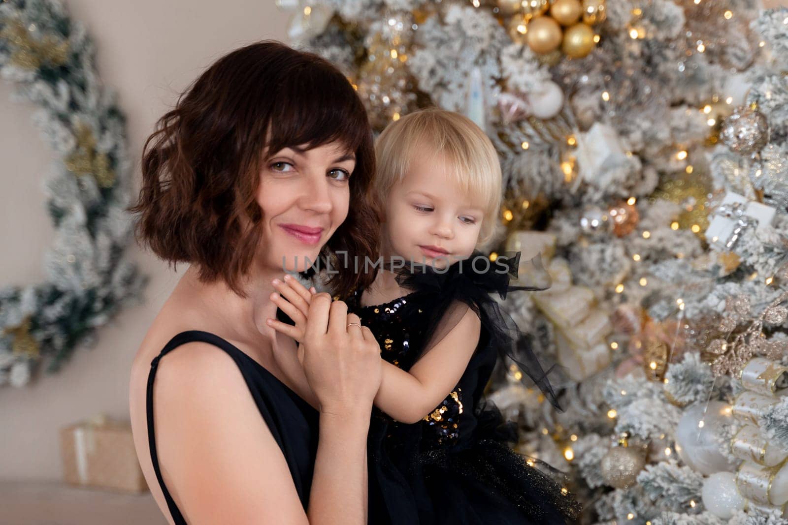 Mother daughter 2 years old Christmas tree. Both are dressed in black dresses, the mother holds the girl in her arms and both look at each other. The family celebrates Christmas