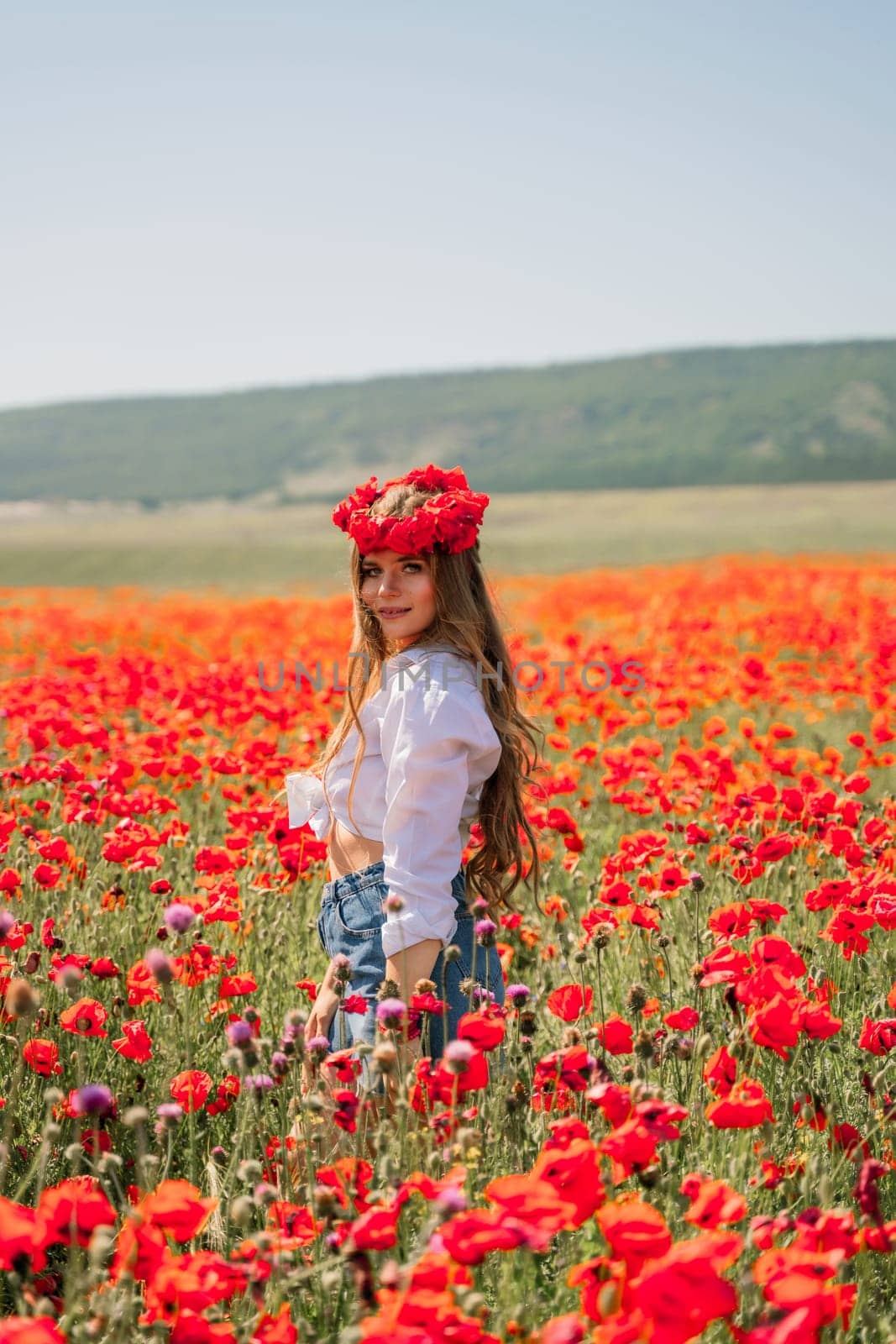 Happy woman in a poppy field in a white shirt and denim skirt with a wreath of poppies on her head posing and enjoying the poppy field