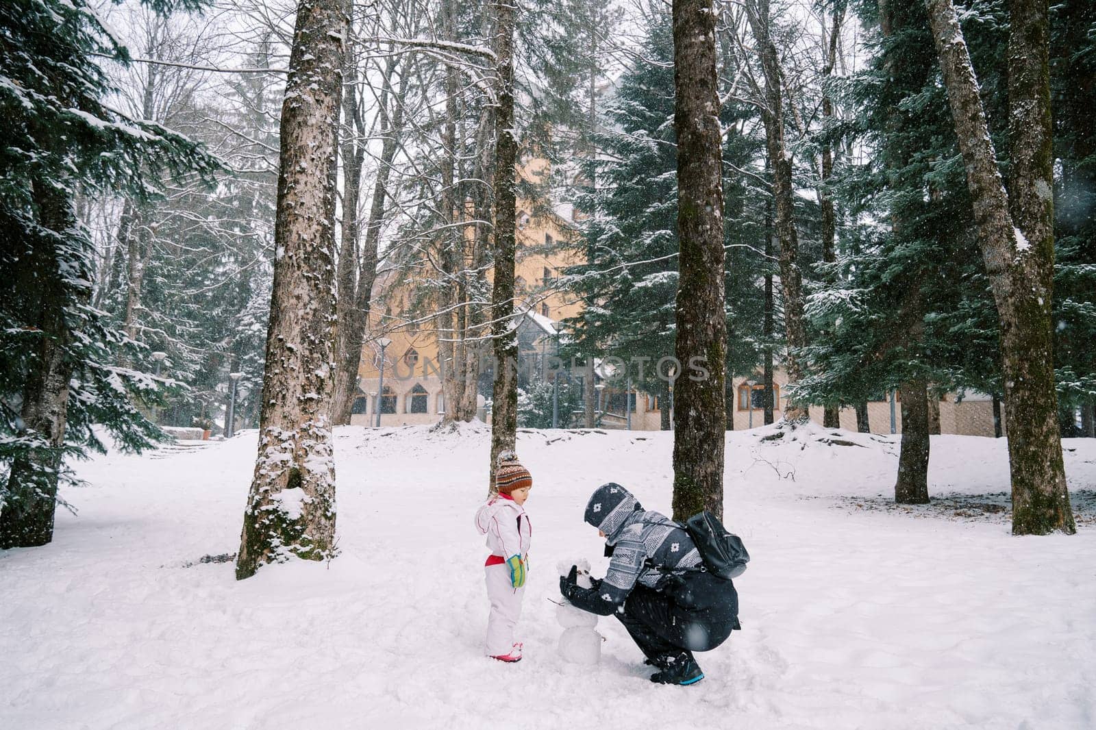 Little girl looks at her mother making a snowman face out of pine cones in the forest. High quality photo
