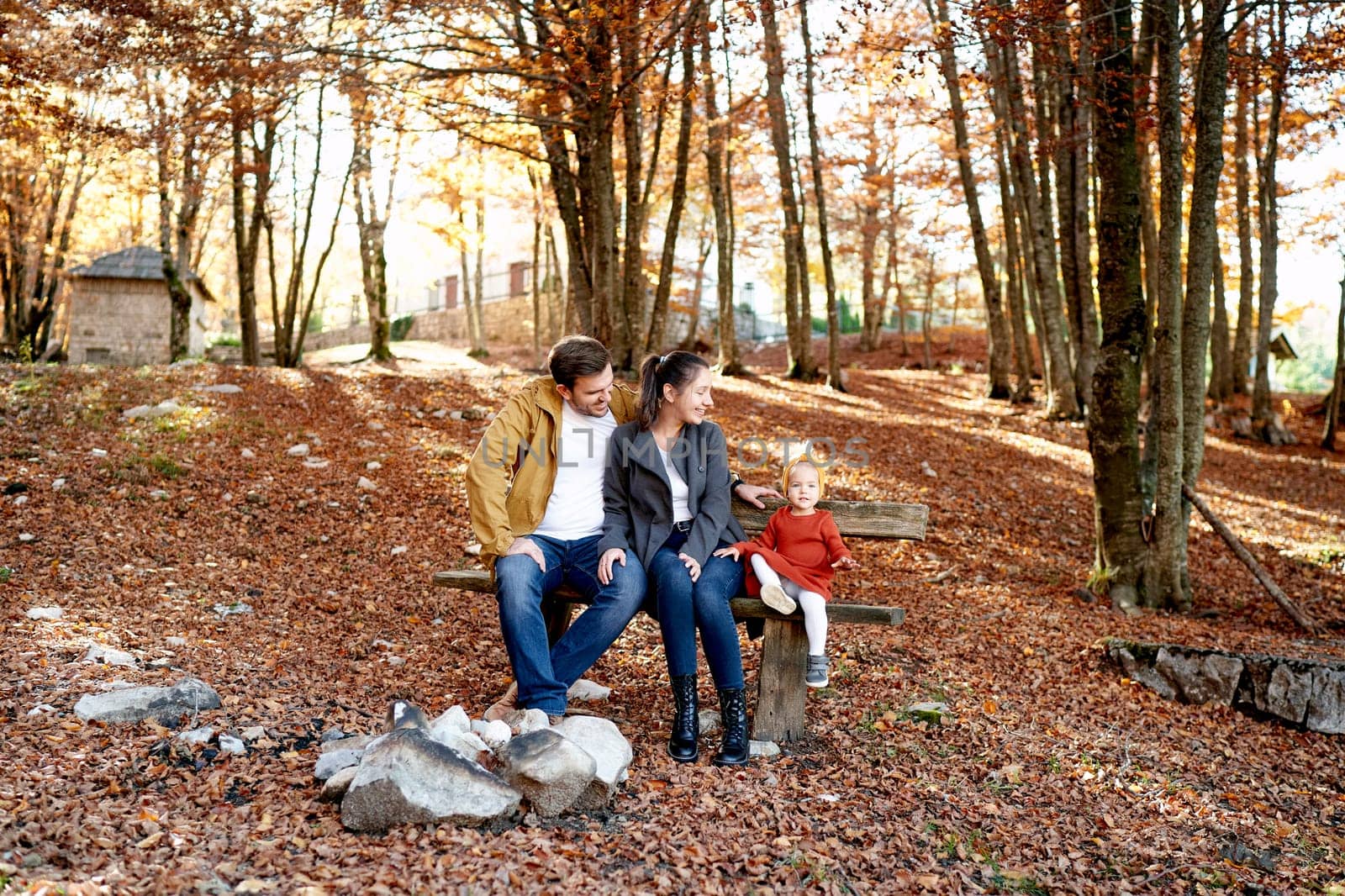 Smiling mom and dad look at a little girl sitting next to her on a bench in the autumn forest. High quality photo