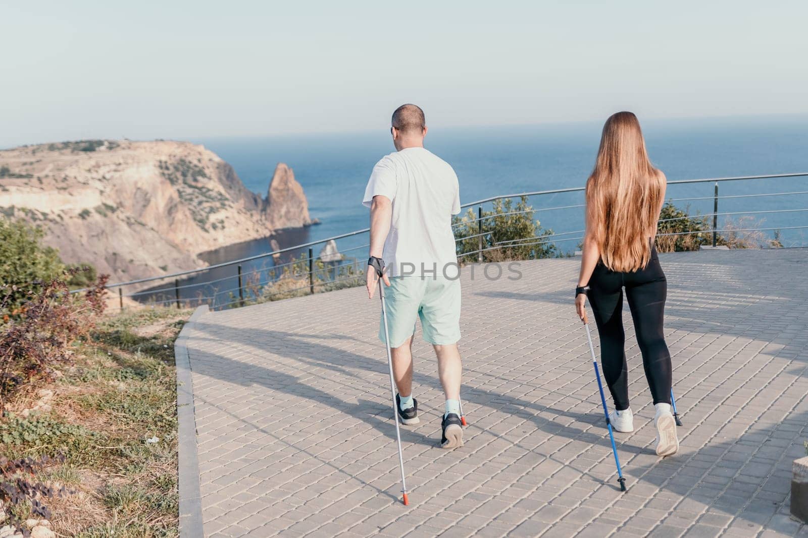 Happy Middle aged couple or friends practicing nordic walking in park near sea. Mature couple with trekking poles walking, practicing Nordic walking outdoors. Aging youthfully and sport concept by panophotograph