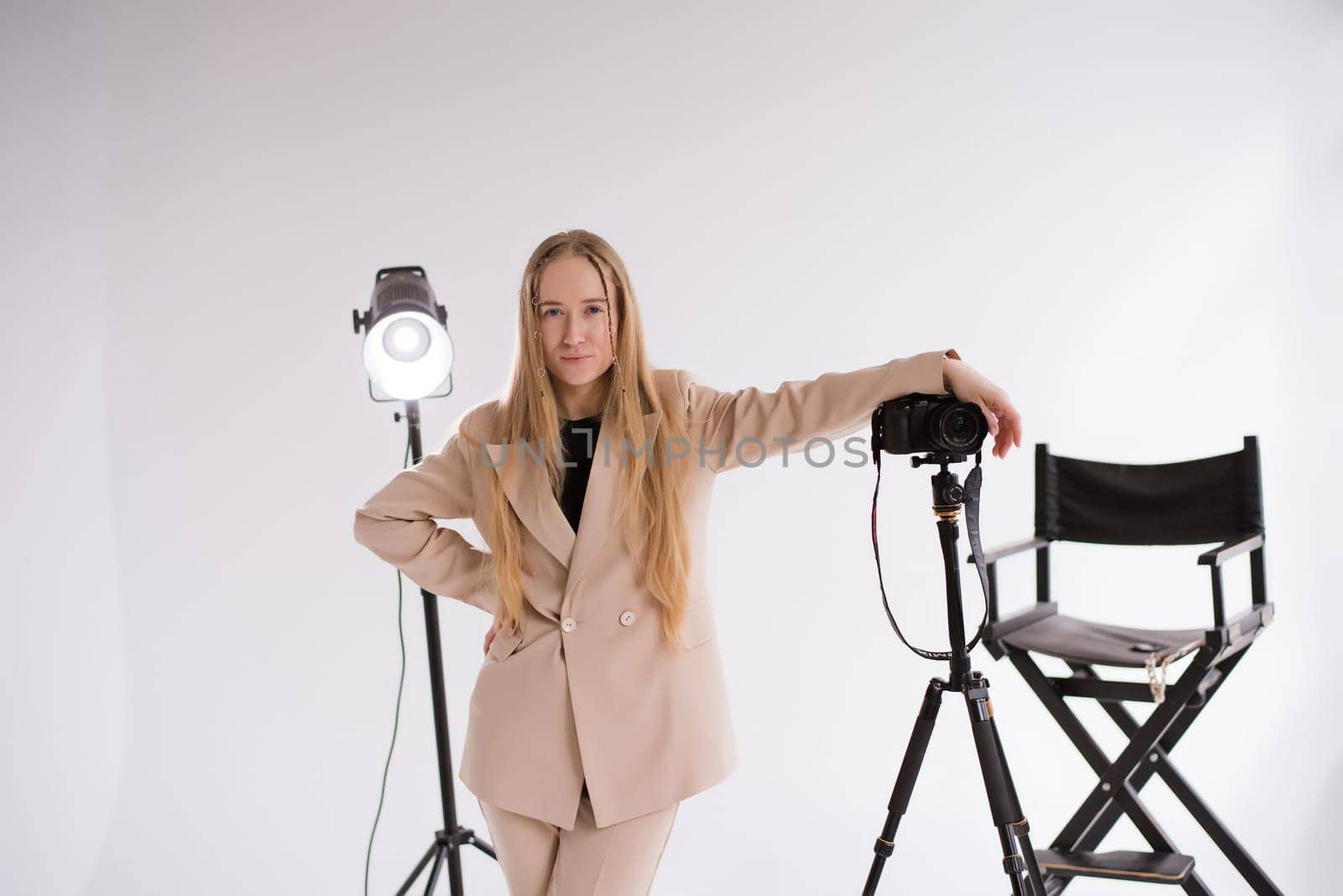 A professional woman producer, videographer, photograph, blogger a blonde working with a camera Lumix GH5, light, tripod in the studio. Wearing a formal nude pantsuit on a white isolated background,