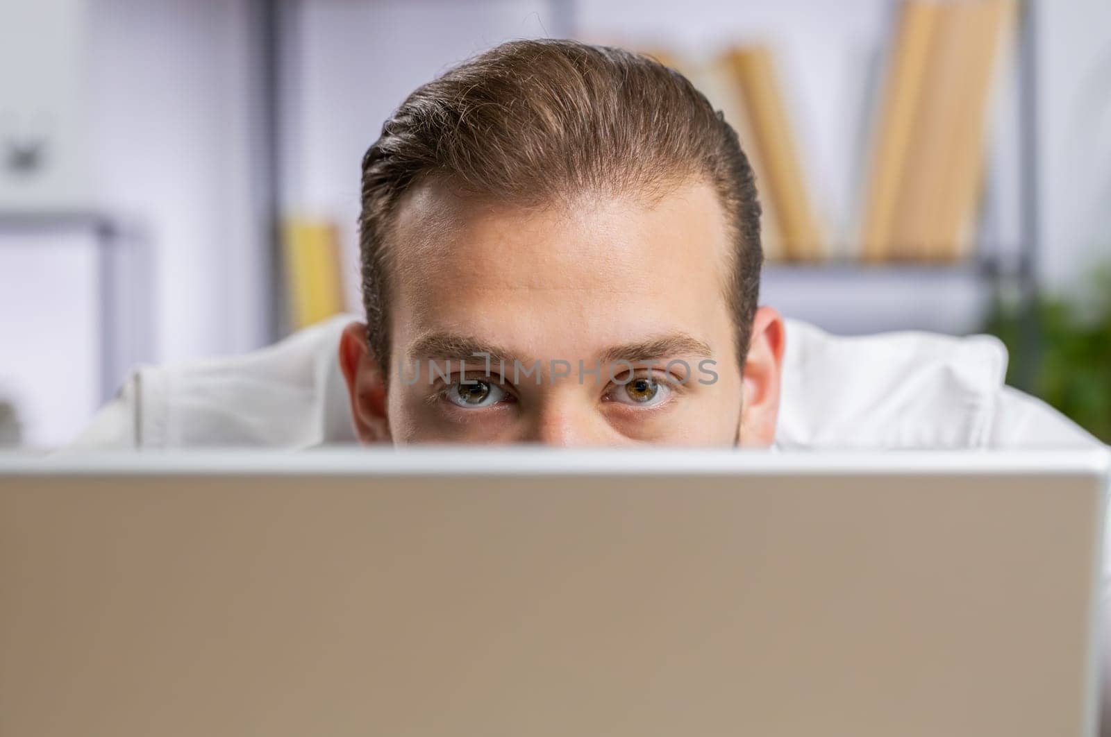 Lebanese confident businessman hiding behind laptop computer, looking at camera, spying his colleagues working, peeping. Professional freelancer man looking from behind computer with cunning eyes
