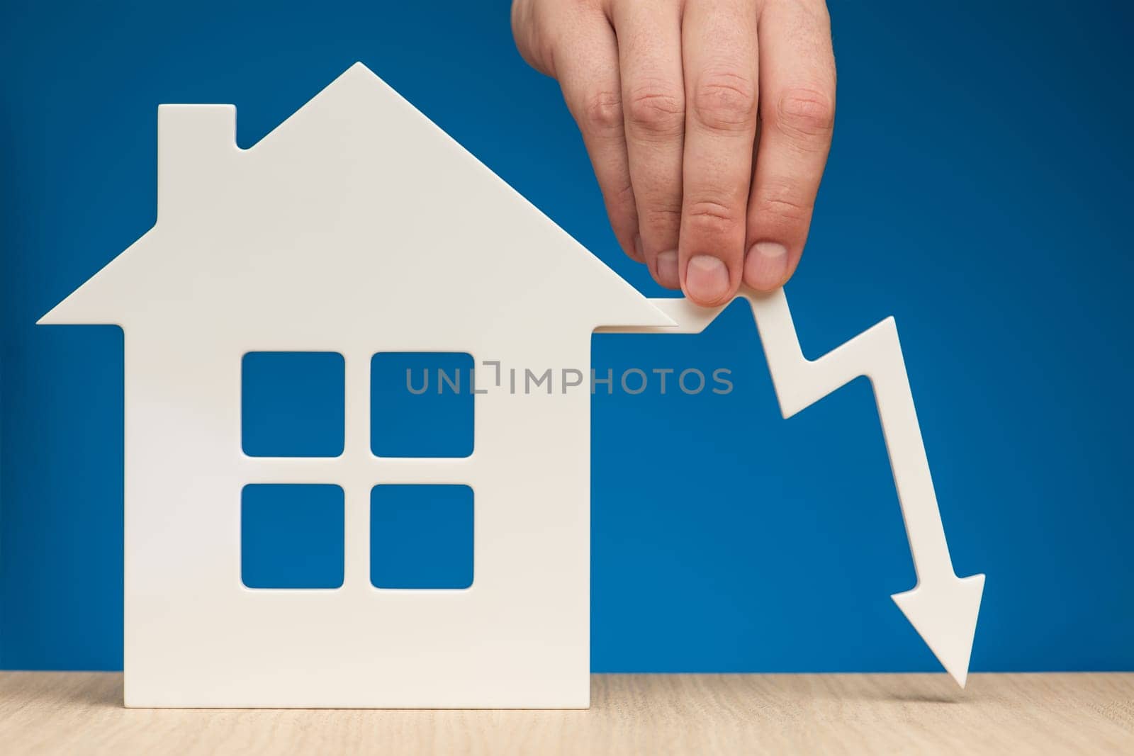 The collapse in real estate prices. Sale in the housing market. Reducing the value of real estate. Model of a house on a blue background and a man's hand holding a white graph arrow pointing down. by SERSOL