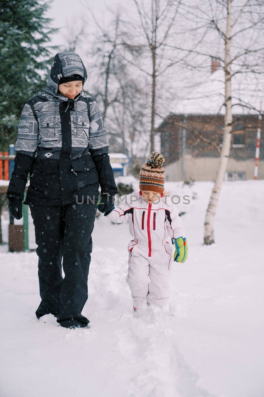 Mom and little girl walk holding hands through the snowdrifts under the snowfall, looking at their feet. High quality photo