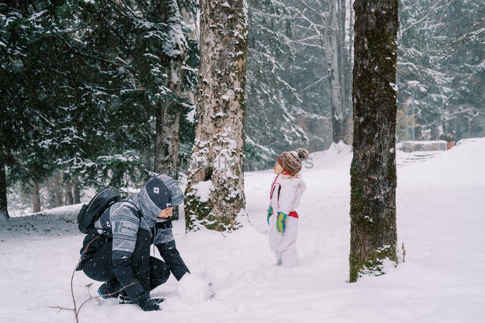 Little girl stands in a snowy forest near her mother making a snowman while squatting by Nadtochiy