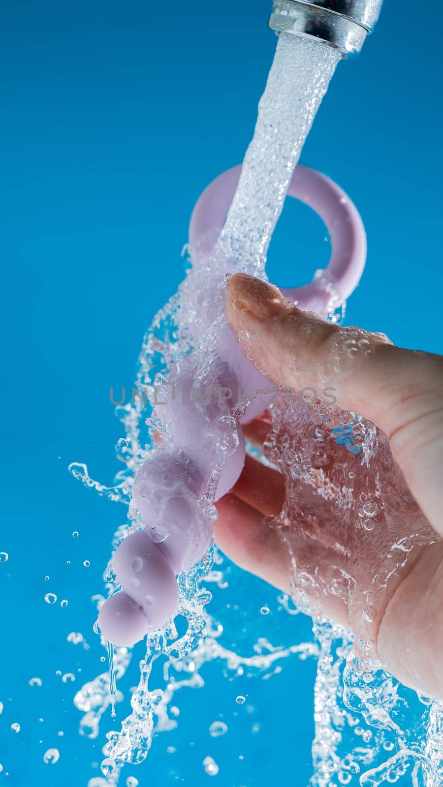 Woman holding lilac anal beads under running water on blue background. Sex toy hygiene concept. by mrwed54