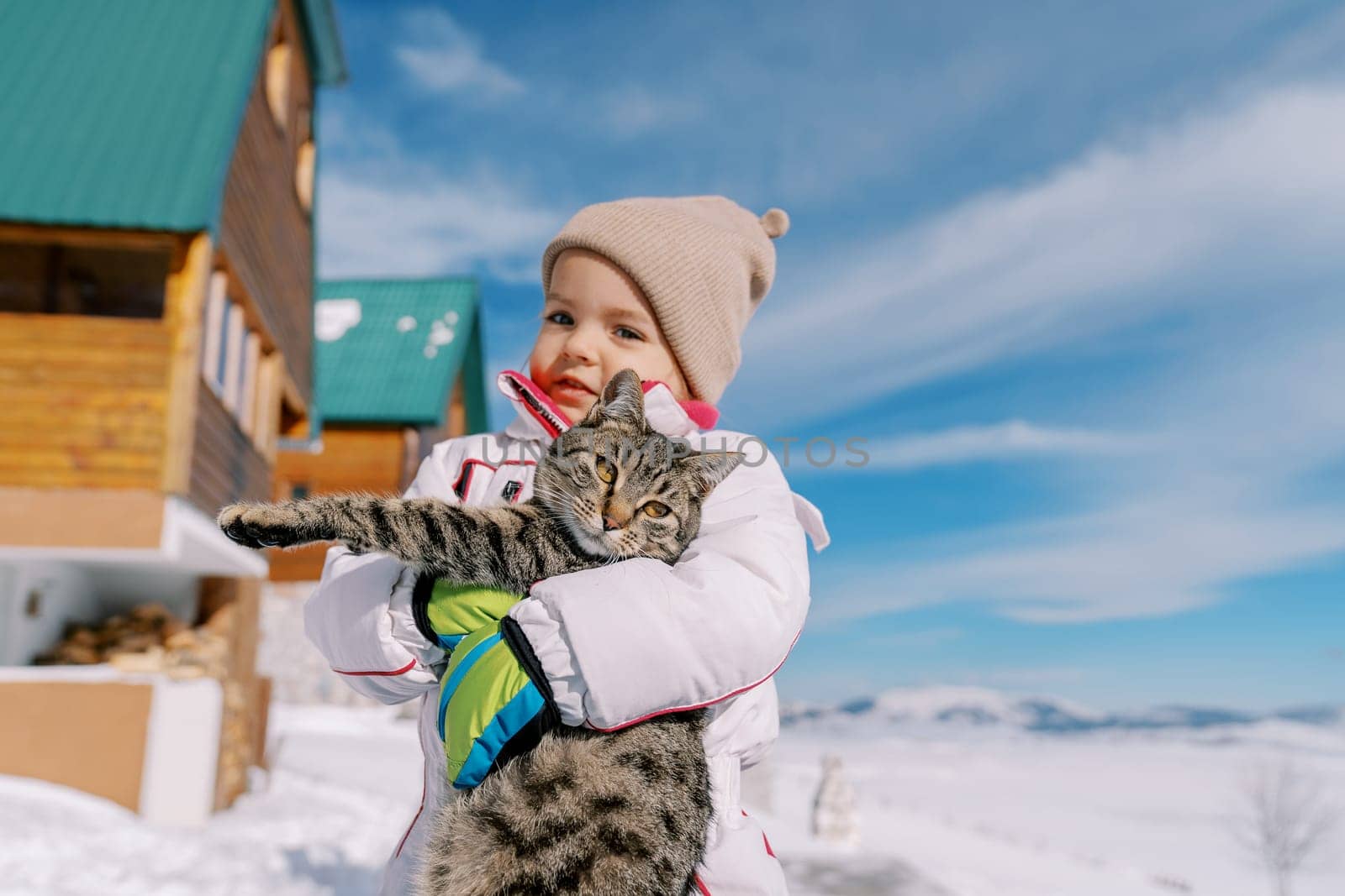 Smiling little girl holding a tabby cat in her arms while standing in the snow near a wooden cottage by Nadtochiy