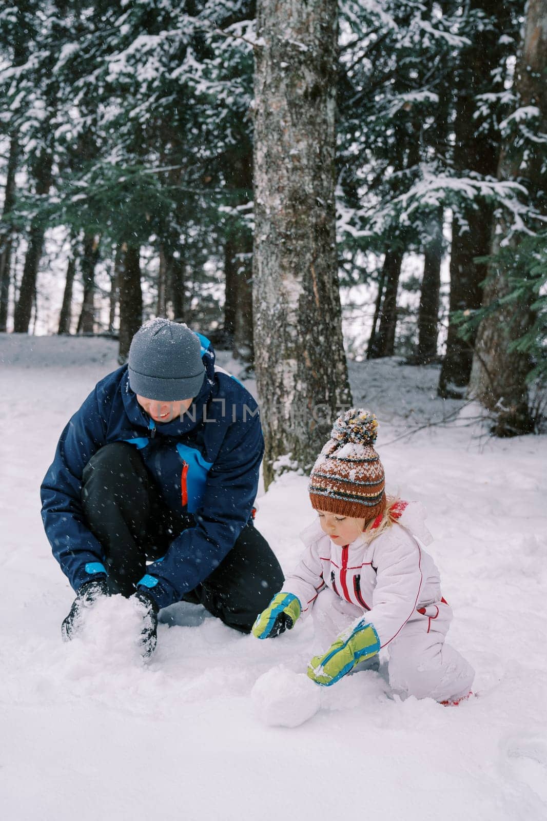 Dad and little girl make a snowman while squatting in a snowy forest by Nadtochiy