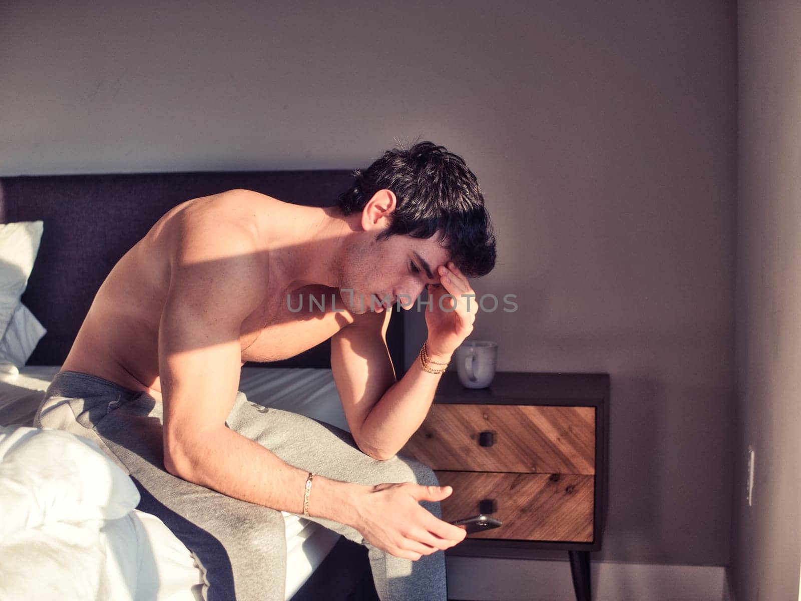 A shirtless man sitting on top of a bed, looking at his cell phone with a worried or sad expression