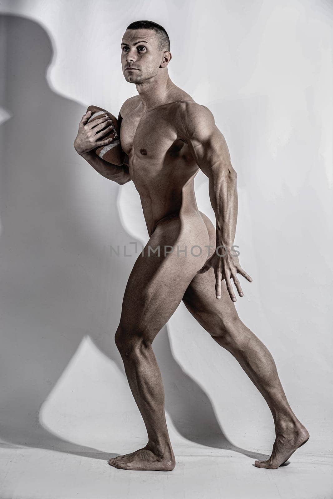 Young muscular naked man in motion of running with football looking concentrated. Full figure shot in studio