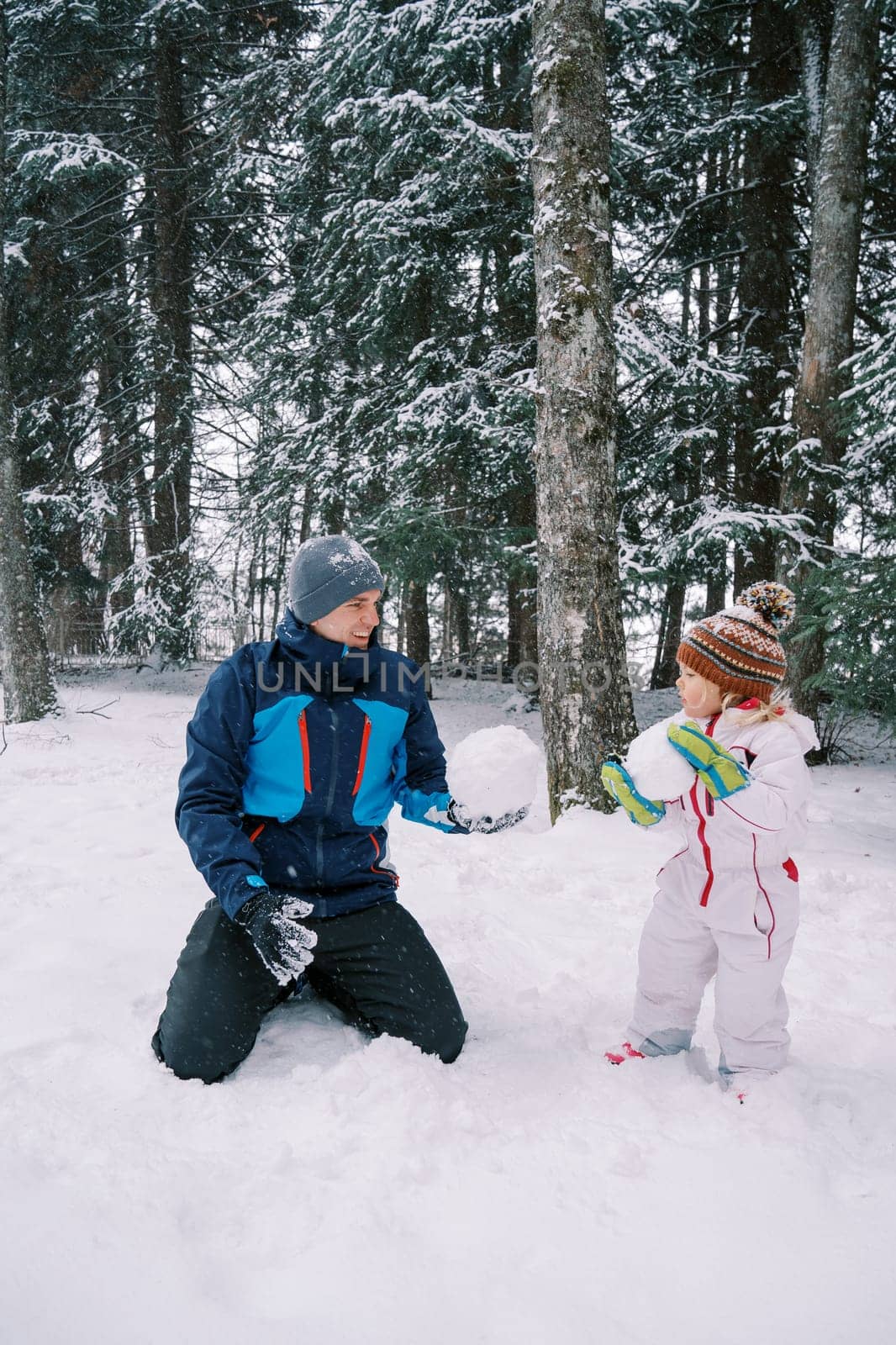 Dad and little girl show each other big snowballs for a snowman in a snowy forest by Nadtochiy