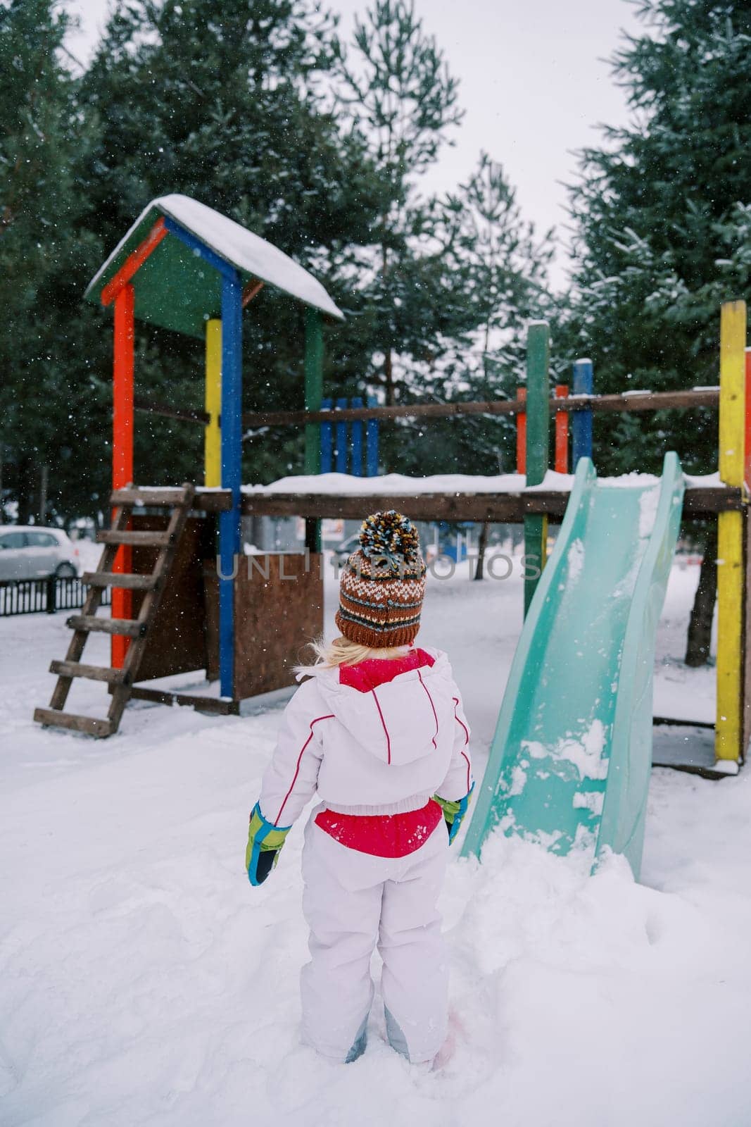 Little girl stands near a snow-covered colorful slide and looks at it. Back view. High quality photo