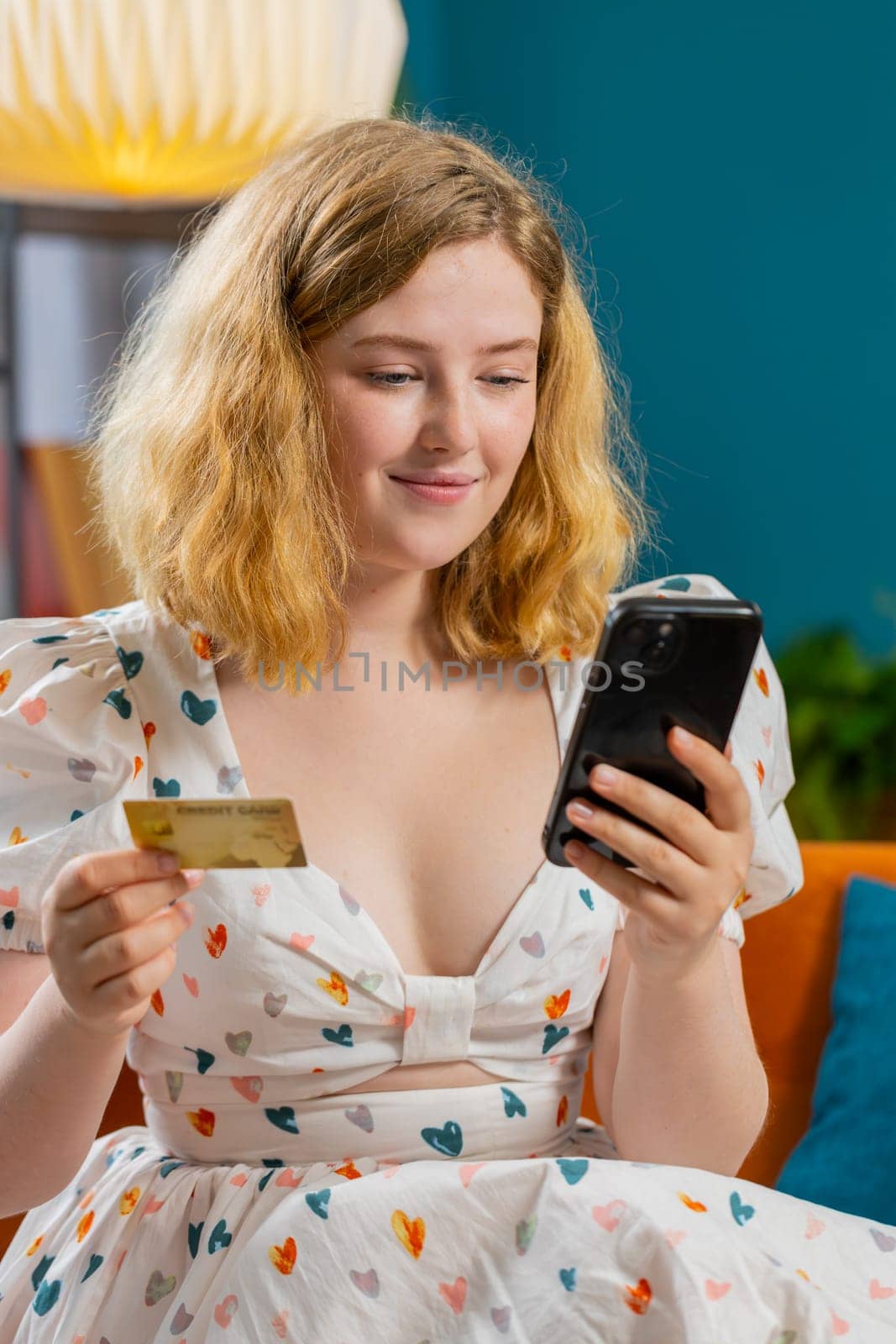 Young redhead woman using credit bank card and smartphone while transferring money, purchases online shopping cashless, order food delivery at home indoors. Happy girl sits in room on couch. Vertical