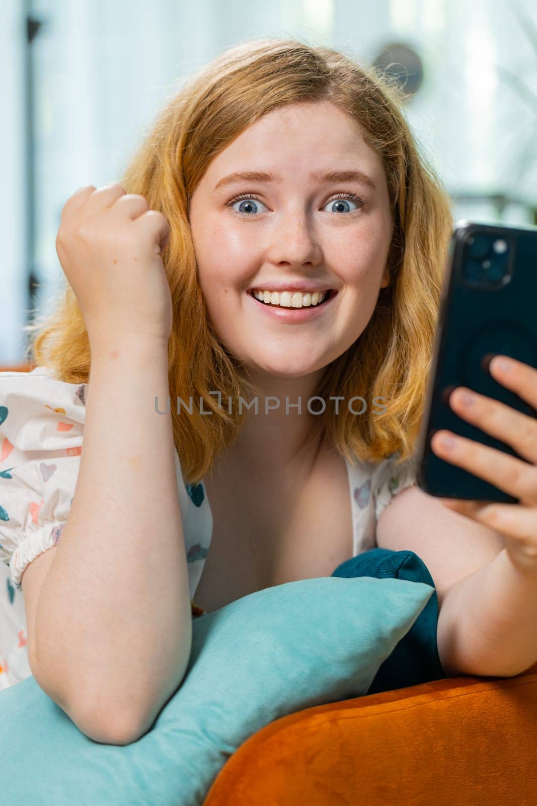 Happy woman use mobile smartphone typing browsing say Wow yes found out great big win good news celebrate lottery jackpot, play victory game. Redhead girl at home in room lying on sofa. Vertical shot