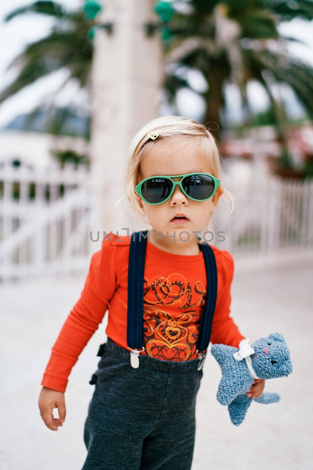 Little girl in sunglasses with a soft toy in her hand stands on the street. High quality photo
