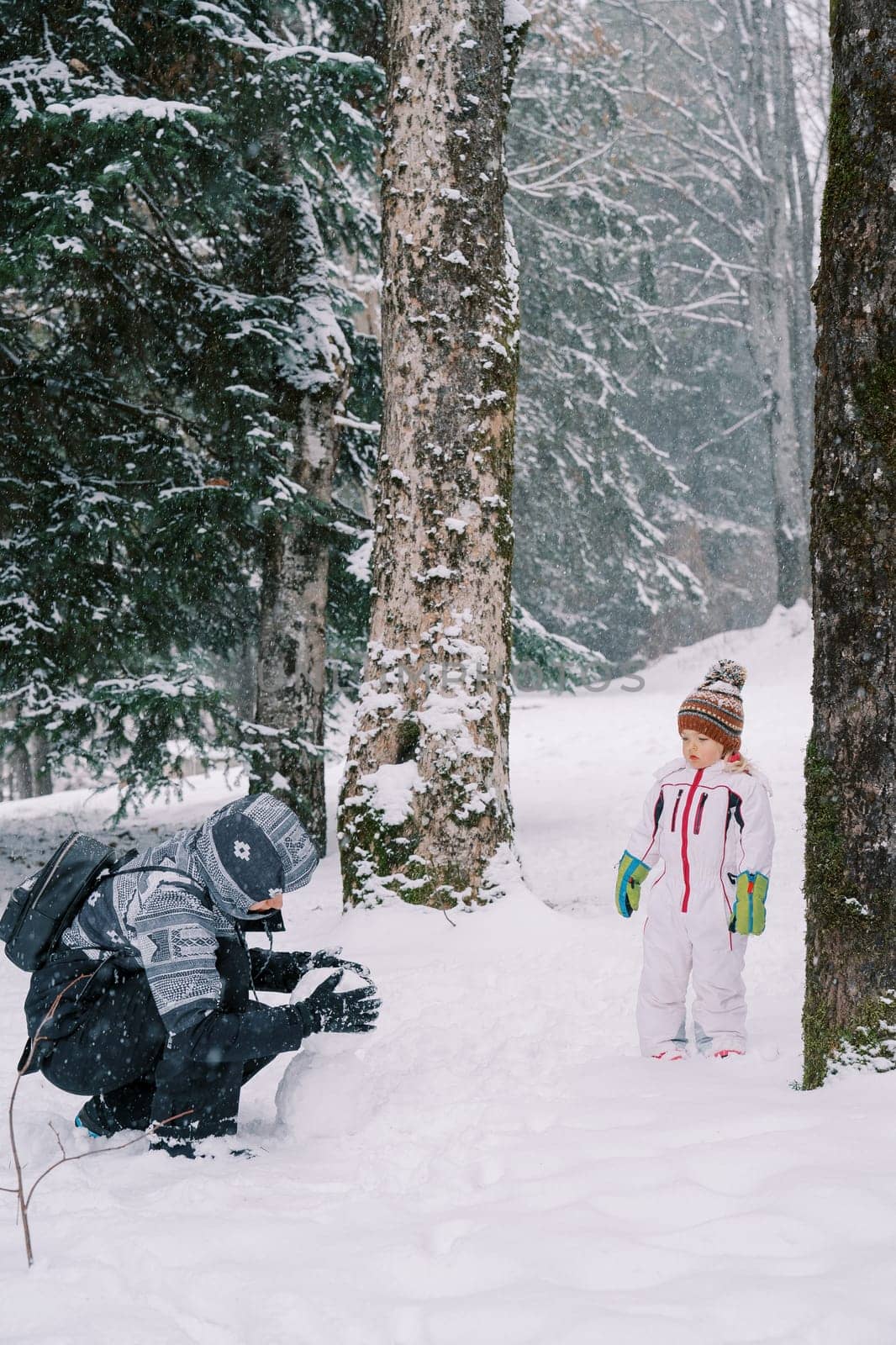 Little girl stands in the forest and watches her mother making a snowman. High quality photo