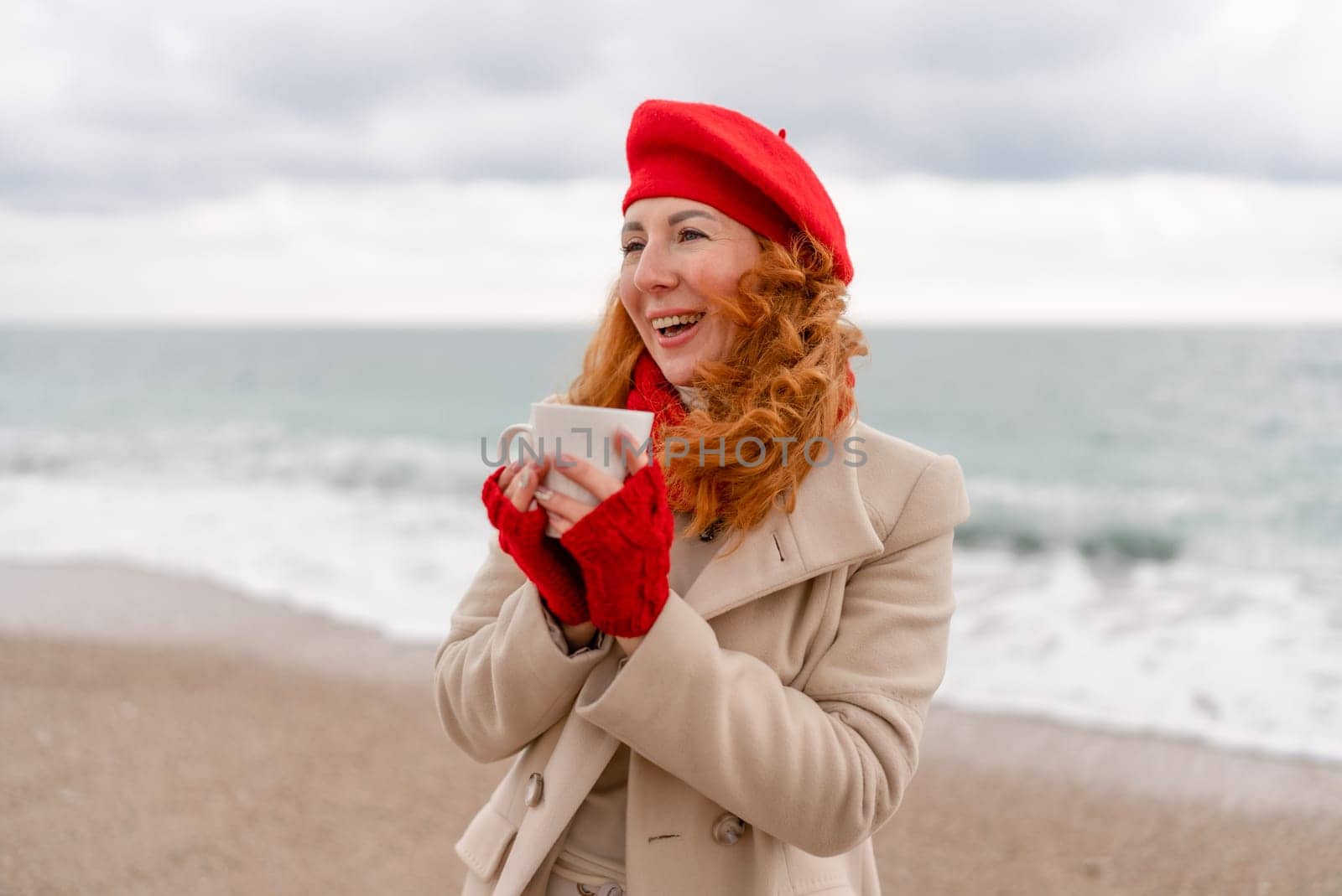 Woman beach in red beret scarf and mitts holding a cup of tea in his hands. Depicting beach relaxation and cozy attire. Walks by the sea.