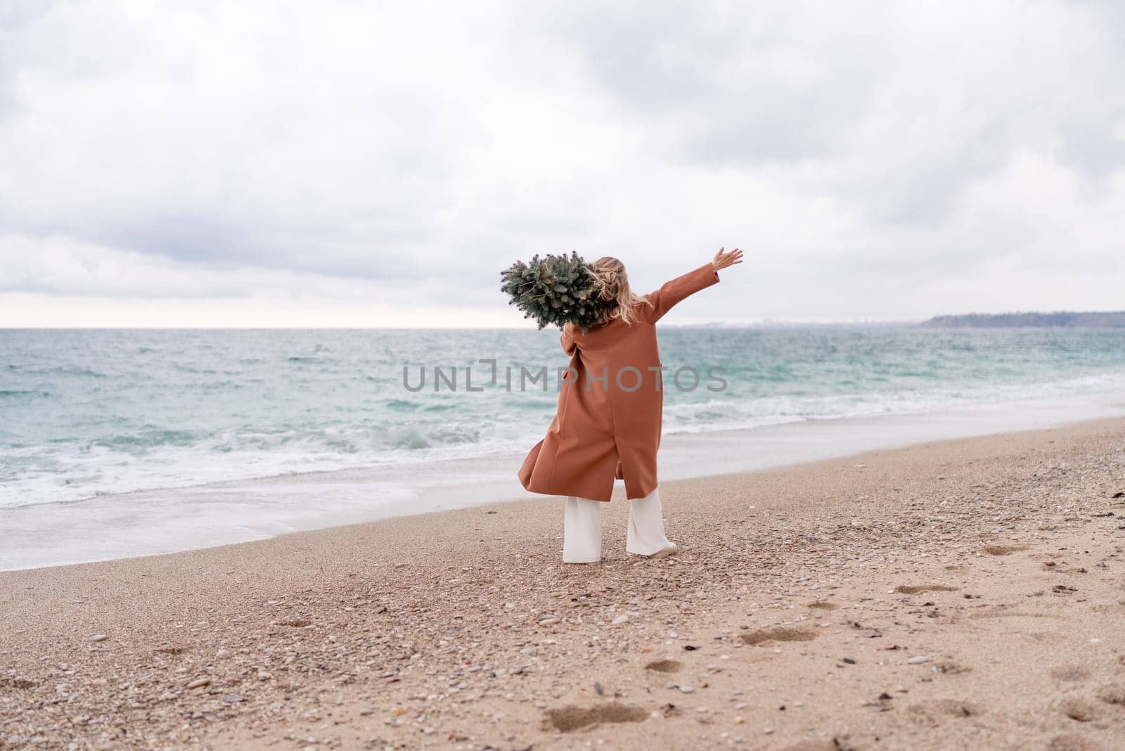 Blond woman Christmas sea. Christmas portrait of a happy woman walking along the beach and holding a Christmas tree on her shoulder. She is wearing a brown coat and a white suit