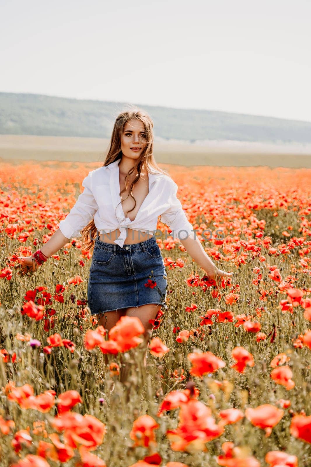 Happy woman in a poppy field in a white shirt and denim skirt with a wreath of poppies on her head posing and enjoying the poppy field. by Matiunina