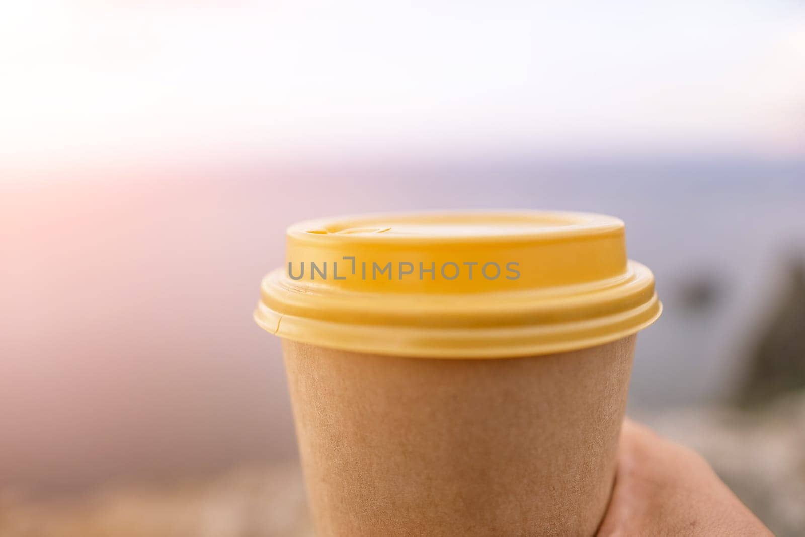 Hand holding Yellow cup with lid, coffee against a backdrop of a blue sky and sea. Illustrating cup and beverage.