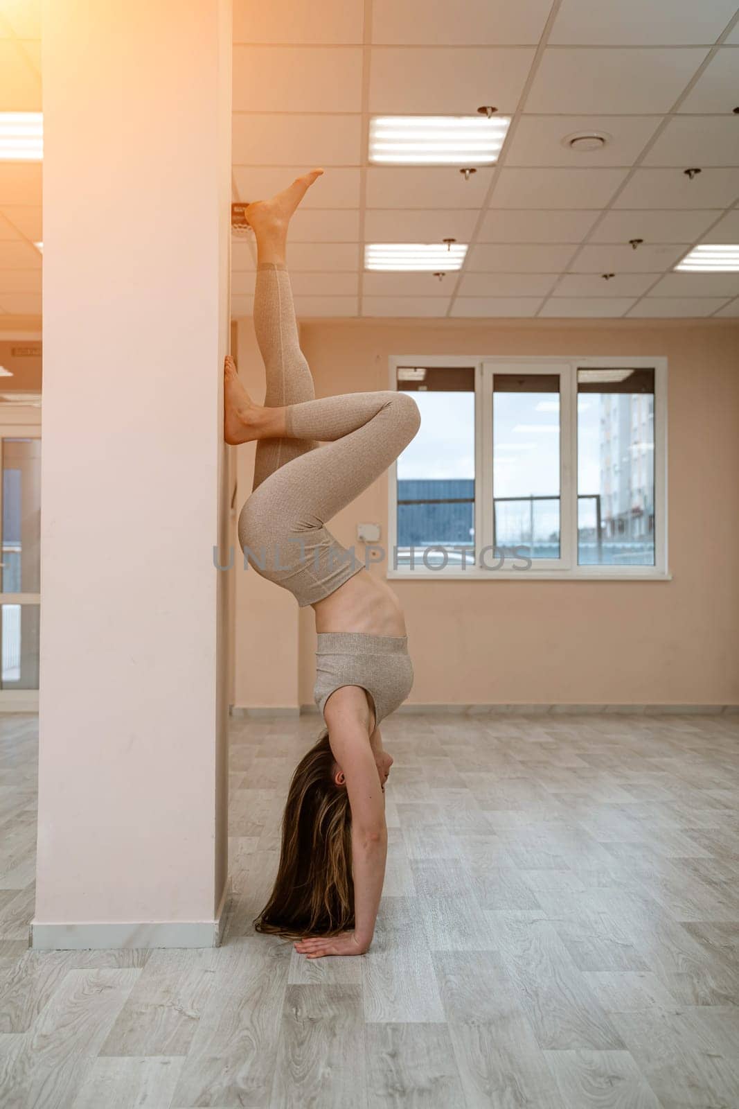 The girl is training in the yoga studio. Dressed in a beige tracksuit doing a handstand against the wall. Healthy lifestyle and yoga concept. by Matiunina