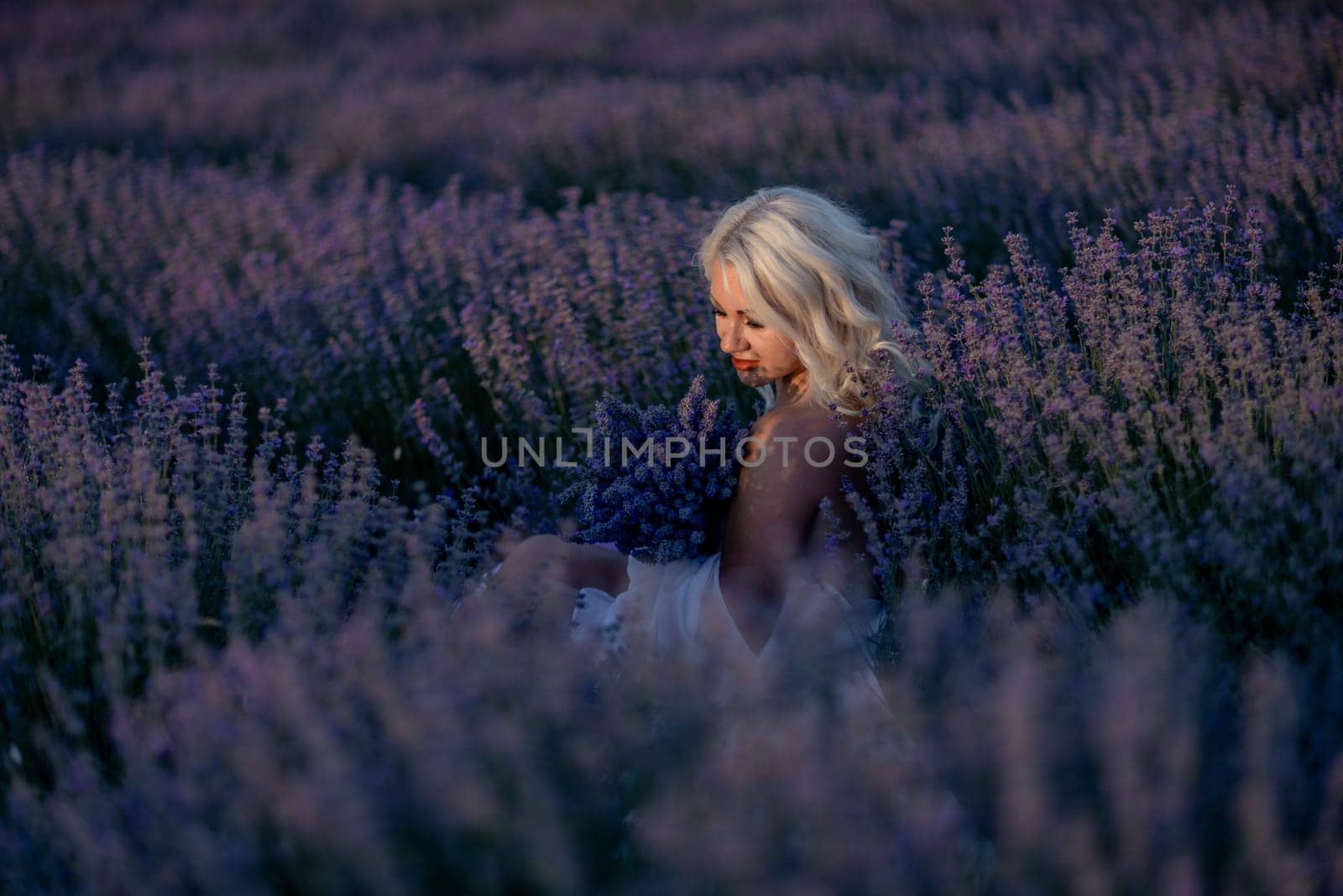 Blonde woman poses in lavender field at sunset. Happy woman in white dress holds lavender bouquet. Aromatherapy concept, lavender oil, photo session in lavender by Matiunina