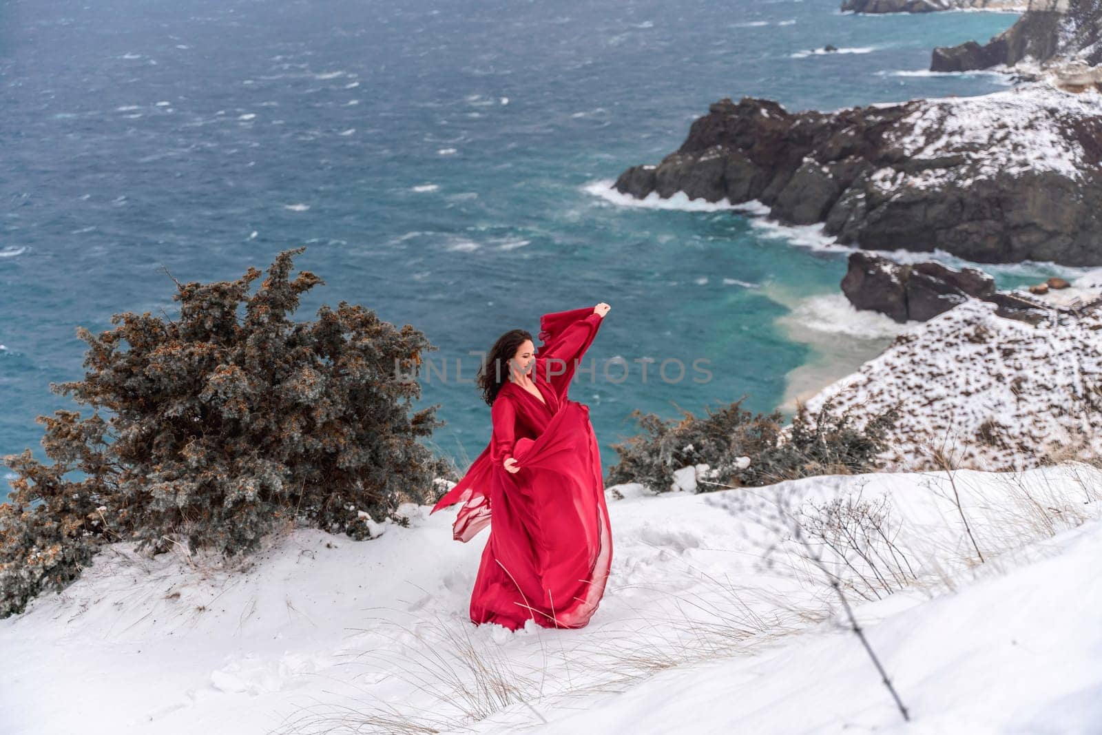 Woman red dress snow sea. Happy woman in a red dress in the snowy mountains by the emerald sea. The wind blows her clothes, posing against sea and snow background. by Matiunina