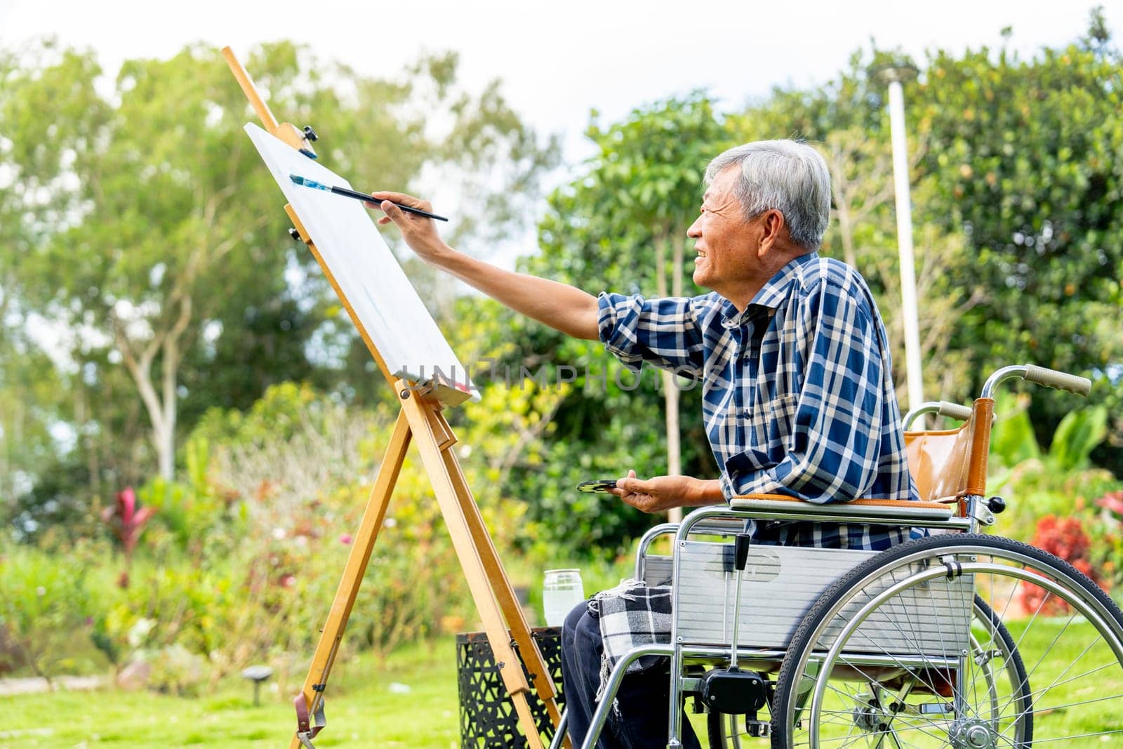 Side view of one Asian senior man sit on wheelchair and paint the painting outdoor for relax and revive good health of old people outside.