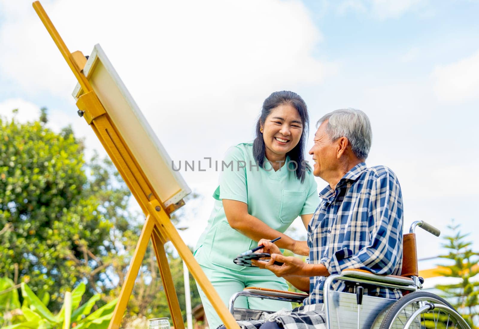 Close up of Asian nurse stand beside senior man sit on wheelchair and draw or paint for relax and they smile and look each other with happiness.