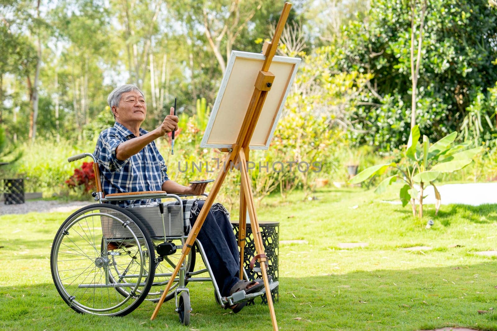 Asian senior man sit on wheelchair and action of aim or point to the area of model for paint the painting outdoor for relax outside.