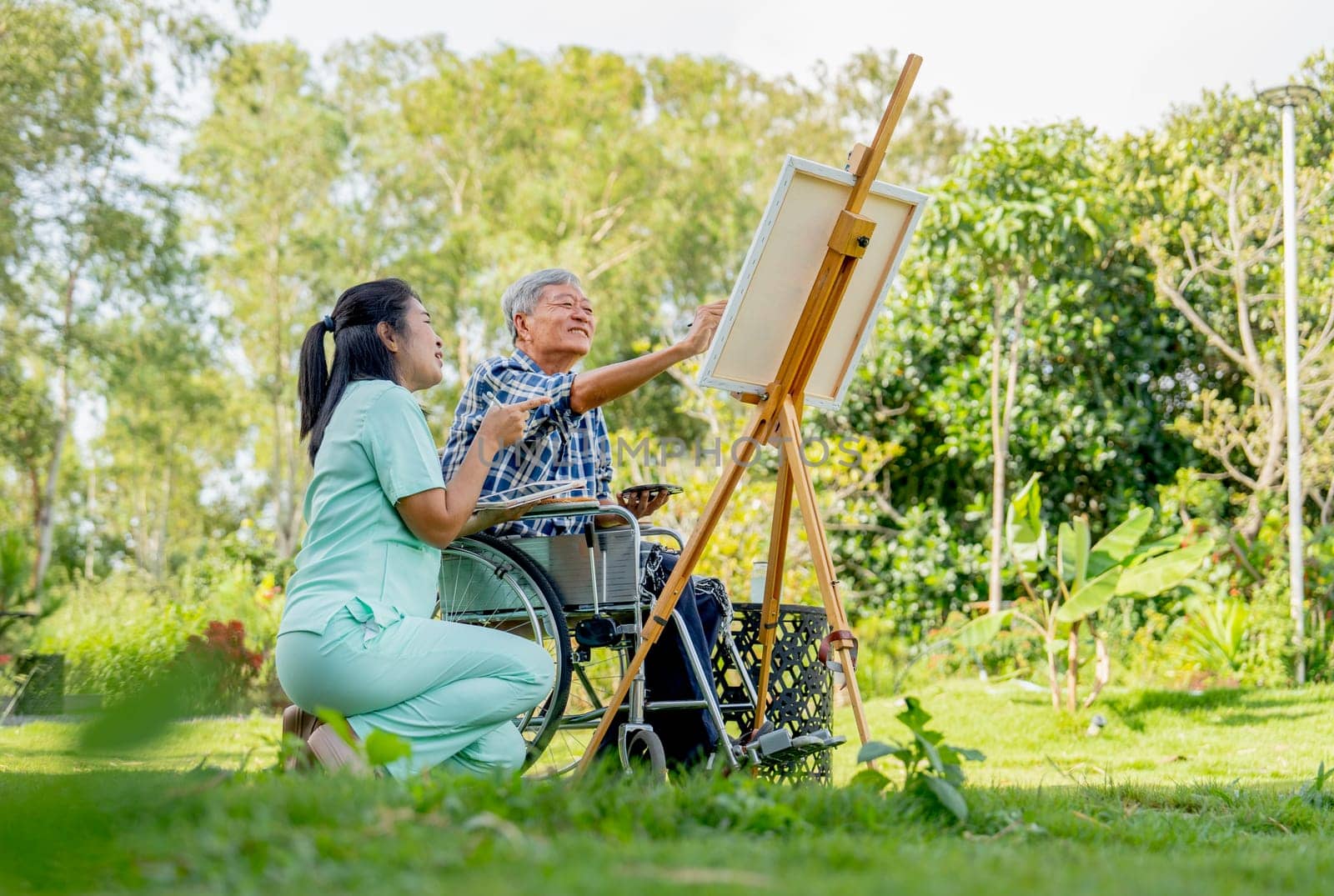Asian nurse woman sit near senior man on wheelchair enjoy to paint the painting outdoor in the garden and they look happiness.